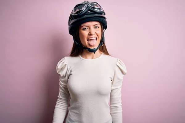 ピンクの背景に元ヘルメットをかぶっている青い目をした若い美しいバイク乗りの女性が 面白い表情で嬉しそうに舌を突き出している 感情概念 — ストック写真