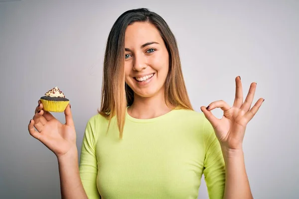 青い目の若い美しい女性は白い背景に甘いチョコレートカップケーキを食べます指でOkサインをします 優れたシンボル — ストック写真