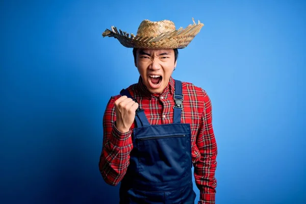 Young handsome chinese farmer man wearing apron and straw hat over blue background angry and mad raising fist frustrated and furious while shouting with anger. Rage and aggressive concept.