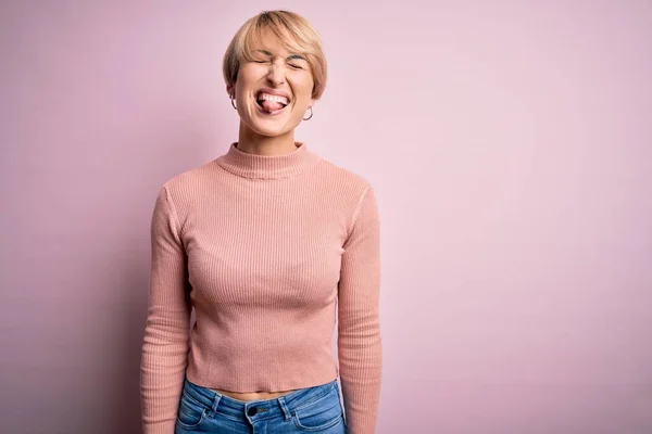 年轻的金发女人 穿着宽松的高领毛衣 粉色背景 带着滑稽的表情伸出舌头 情感概念 — 图库照片
