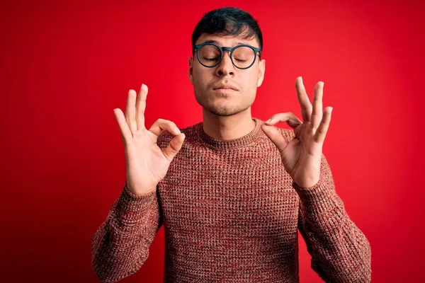 赤を背景にオタク眼鏡をかけた若いハンサムなヒスパニック系男性がリラックスし 指で瞑想のジェスチャーを行う目で笑顔を閉じました ヨガのコンセプト — ストック写真
