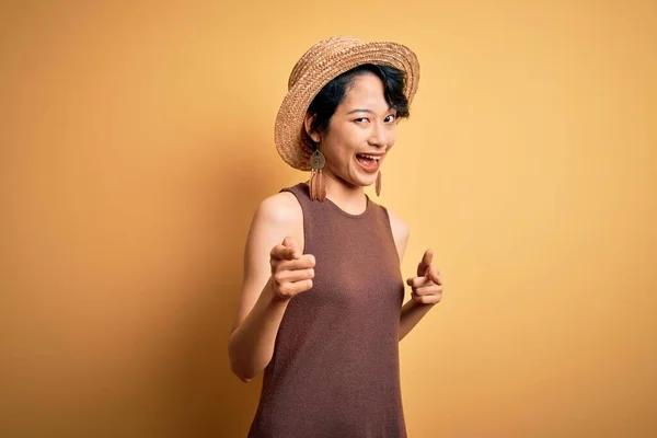若い美しいアジアの女の子が幸せと面白い顔を持つカメラに指を指して 孤立した黄色の背景の上にカジュアルなTシャツと帽子を着て 良いエネルギーと振動 — ストック写真