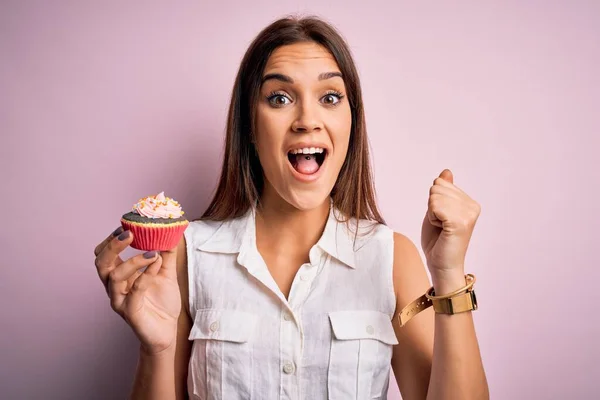 若い美しいブルネットの女性食べるチョコレートカップケーキ以上隔離されたピンクの背景叫び誇りと勝利と成功を祝う非常に興奮し 応援感情 — ストック写真