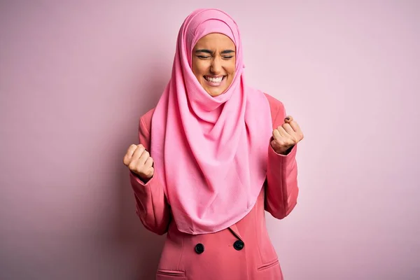 ピンクのイスラム教徒のヒジャーブとビジネスジャケットを身に着けている若い美しいブルネットの実業家は非常に幸せと腕を上げて勝者のジェスチャーをやって興奮し 笑顔と成功のために叫んでいます お祝いのコンセプト — ストック写真
