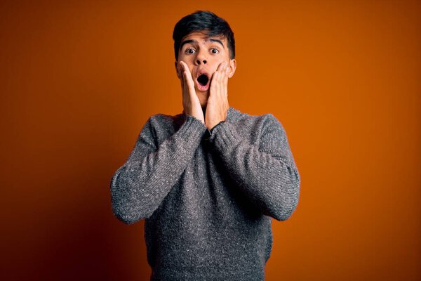Young handsome man wearing casual sweater standing over isolated orange background afraid and shocked, surprise and amazed expression with hands on face