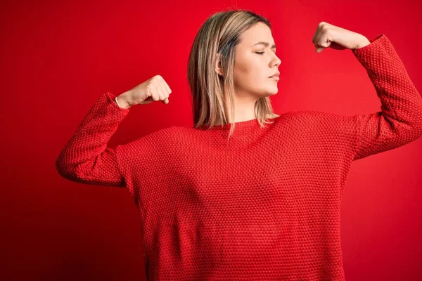 Young beautiful blonde woman wearing casual sweater over red isolated background showing arms muscles smiling proud. Fitness concept.