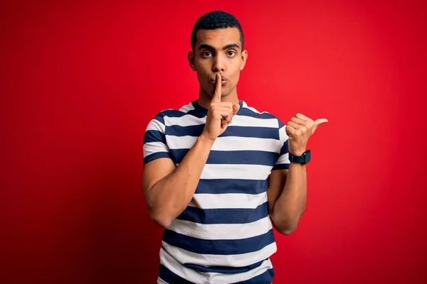 英俊的非洲裔美国男人穿着休闲条纹T恤 站在红色的背景上 要求安静 手指放在嘴唇上 手拉手指向侧面 沉默和秘密概念 — 图库照片