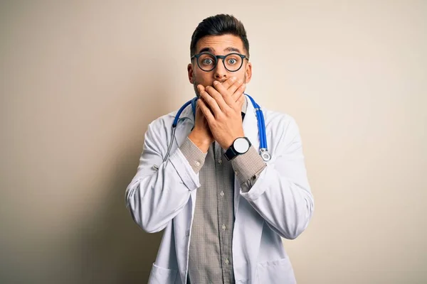 眼鏡をかけた若い医者の男性 隔離された背景の上の医学の白いローブそして聴診器は間違いのための手で口を覆う衝撃を与えた 秘密概念 — ストック写真
