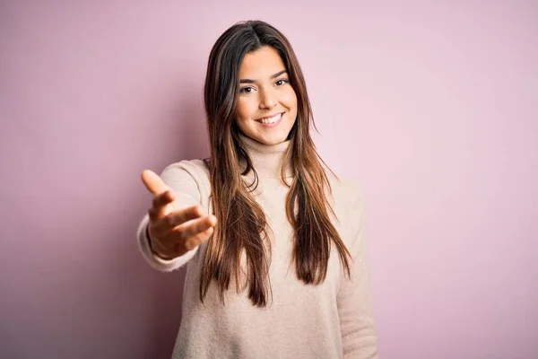 年轻美丽的女孩穿着宽松的高领毛衣 站在孤立的粉色背景上 微笑友好地握手致意和欢迎 成功的业务 — 图库照片