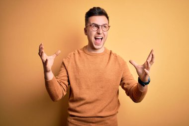 Gözlük takan yakışıklı beyaz bir adam ve sarı arka plan üzerine sıradan bir kazak çılgın ve sinirli bir şekilde bağırıyor ve agresif bir ifade ve kolla bağırıyor. Hayal kırıklığı konsepti.