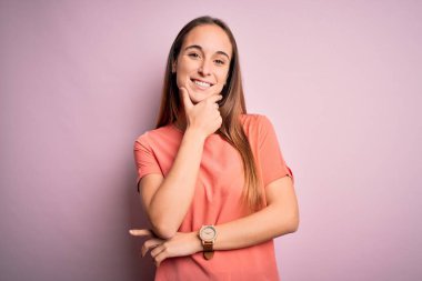 Izole pembe arka plan üzerinde duran rahat t-shirt giyen genç güzel kadın çapraz kollar ve çene üzerinde kaldırdı el ile gülümseyerek kamera emin görünümlü. Olumlu düşünme.