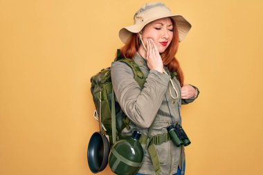 Sırt çantası ve sarı arka planda şapka takan genç kızıl saçlı kadın diş ağrısı ya da diş ağrısı yüzünden ağzıyla dokunuyor. Dişçi