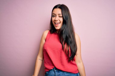 Genç esmer kadın pembe izole edilmiş arka plan üzerine gündelik bir tişört giyip kameraya seksi bir ifadeyle bakıyor, neşeli ve mutlu bir yüzle..