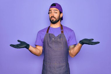 Profesyonel önlük giyen yakışıklı İspanyol bir adam dövme sanatçısı olarak çalışıyor. Kolları ve elleri havada, kafası karışmış bir ifade takınıyor. Şüphe kavramı.