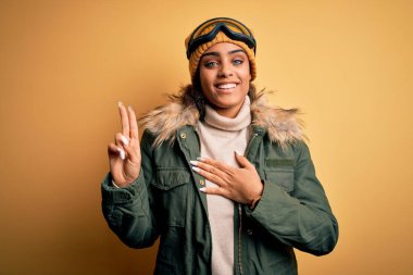 Genç Afrikalı Afro-Amerikan kayakçı kız kar sporu kıyafetleri ve kayak gözlükleriyle gülümsüyor. Göğüs ve parmaklar havada küfrederek sadakat yemini ediyor.