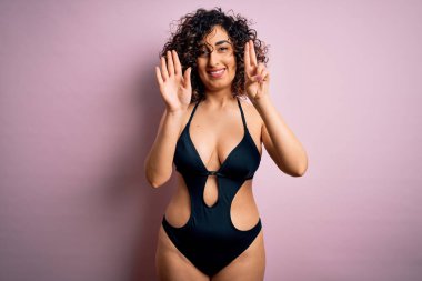 Tatilde olan genç, güzel bir Arap kadın. Pembe arka planda mayo ve güneş gözlüğü takıyor. Yedi numarayı işaret ediyor ve gülümsüyor..