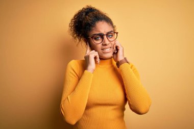 Sarı arka plan üzerine süveter ve gözlük takan Afro-Amerikan genç bir kız yüksek sesli müzik sesi için kulaklarını parmaklarıyla kapatıyor. Sağır kavramı.