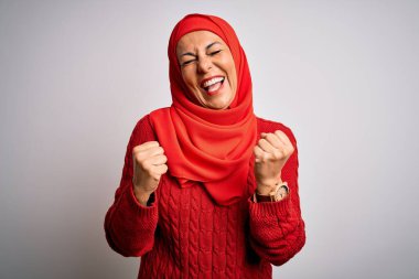 Orta yaşlı esmer kadın izole edilmiş beyaz arka plan üzerine geleneksel tesettür takmış kolları havada ve gözleri kapalı bir şekilde başarıyı kutluyor ve şaşırıyor. Kazanan konsept.