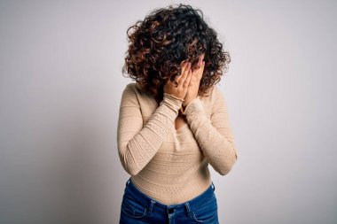 Genç, kıvırcık, güzel bir Arap kadın beyaz arka planda günlük tişört ve gözlük takıyor. Ağlarken suratını ellerle kapatıyor. Depresyon kavramı.