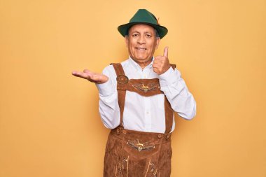 Sarı arka planda Alman geleneksel ahşap şenliği kıyafeti giyen yaşlı gri saçlı adam avuç içini gösteriyor ve baş parmaklarıyla iyi bir hareket yapıyor, mutlu ve neşeli gülümsüyor.