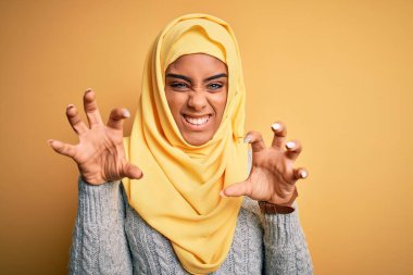 Genç ve güzel Afro-Amerikalı kız izole edilmiş sarı arka plan üzerine Müslüman başörtüsü takıyor komik bir şekilde kedi gibi pençe hareketi yapıyor, agresif ve seksi bir ifade.