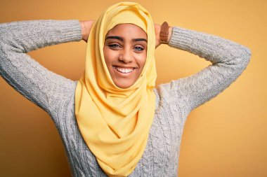 Genç ve güzel Afro-Amerikalı kız izole edilmiş sarı arka plan üzerine Müslüman başörtüsü takıyor rahatlıyor ve geriniyor, kolları ve elleri başının ve boynunun arkasında mutlu bir şekilde gülümsüyor.