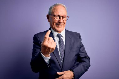 Gri saçlı, gözlüklü, şık takım elbiseli ve mor arka planda kravat takan kıdemli iş adamı. Buraya gel ve mutlu bir şekilde karşıla ve gülümse.