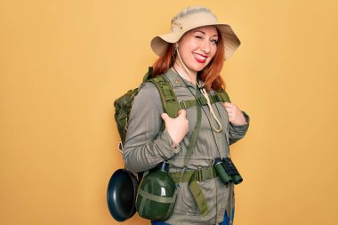 Sırt çantası ve sarı arka planda şapka takan genç kızıl saçlı kadın kameraya seksi bir ifadeyle bakıyor, neşeli ve mutlu bir yüzle..