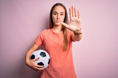 Genç ve güzel bir kadın futbol topunu pembe arka planda tutarak oynuyor. Açık elle dur işareti yapıyor. Ciddi ve kendinden emin bir ifadeyle, savunma jesti.