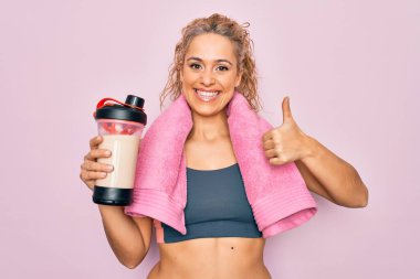 Güzel, sarışın, spor yapan, havluyla protein içeceği sallayan, mutlu ve pozitif gülümseyen, başparmağını kaldıran, mükemmel ve onay işareti yapan bir kadın.