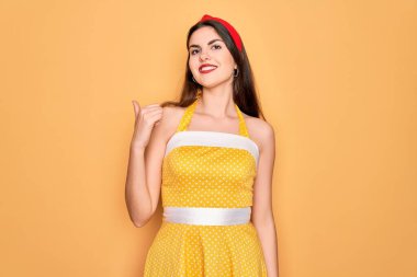 50 'li yılların modasına uygun, sarı arka planda giyinmiş, gülümseyen, gülümseyen ve baş parmağıyla yüzünü gösteren genç ve güzel bir kadın..