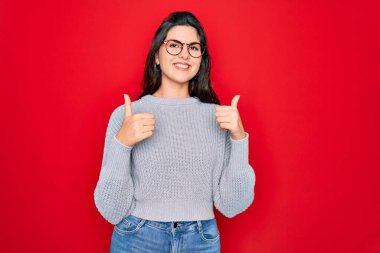 Genç, güzel, esmer bir kadın kırmızı arka plan başarı tabelasının üzerine günlük süveter giyiyor. Elleriyle olumlu jest yapıyor, gülümsüyor ve mutlu. Neşeli ifade ve kazanan jest.