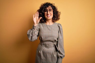 Sarı arka plan üzerinde duran elbise ve gözlük takan güzel Arap iş kadını beş numaralı parmaklarıyla işaret ederken kendinden emin ve mutlu bir şekilde gülümsüyor..