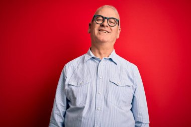 Orta yaşlı, yakışıklı, beyaz, sıradan çizgili gömlek giyen ve kırmızı arka planda gözlüklü, yüzünde mutlu ve havalı bir gülümseme olan bir adam. Şanslı kişi..