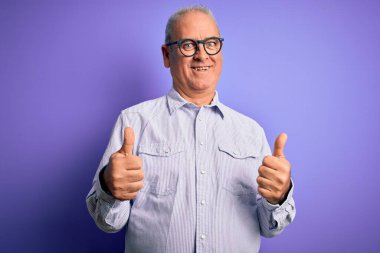 Orta yaşlı, beyaz, çizgili gömlek ve gözlük takan yakışıklı bir adam mor arka plan başarısı tabelasının üzerine el hareketleriyle olumlu bir hareket yapıyor, gülümsüyor ve mutlu. Neşeli ifade ve kazanan jest.