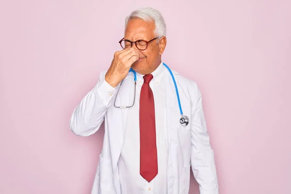 Middle age senior grey-haired doctor man wearing stethoscope and professional medical coat smelling something stinky and disgusting, intolerable smell, holding breath with fingers on nose. Bad smell
