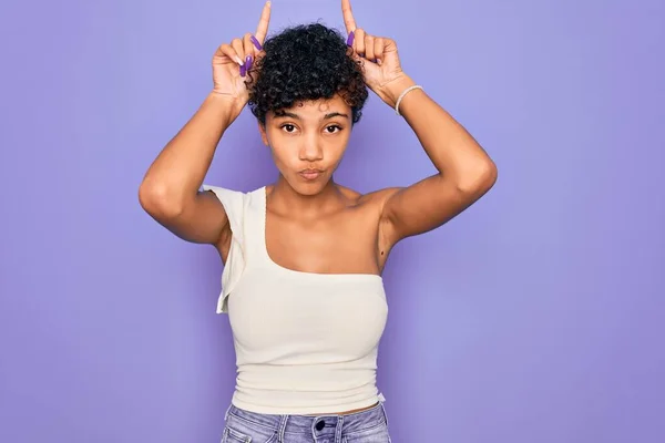 若いです美しいですアフリカ系アメリカ人アフロ女性身に着けていますカジュアルTシャツ以上紫の背景を行います面白いジェスチャーとともに指以上頭としてブル角 — ストック写真