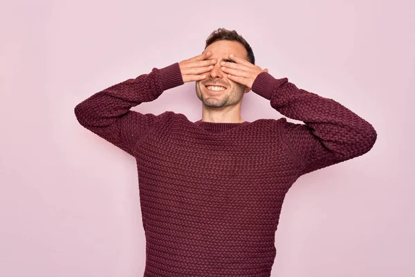 Young handsome man with blue eyes wearing casual sweater standing over pink background covering eyes with hands smiling cheerful and funny. Blind concept.