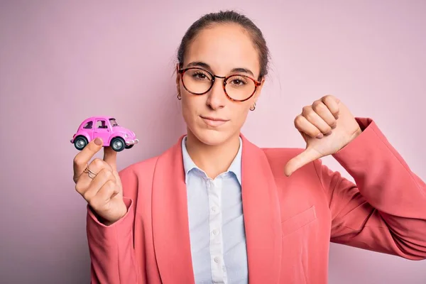 ピンク色の車のおもちゃを持っているメガネを身に着けている若い美しい実業家怒っている顔で隔離された背景に 親指で嫌いを示す負の記号ダウン 拒絶概念 — ストック写真