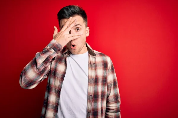 Young handsome caucasian man wearing casual modern shirt over red isolated background peeking in shock covering face and eyes with hand, looking through fingers with embarrassed expression.