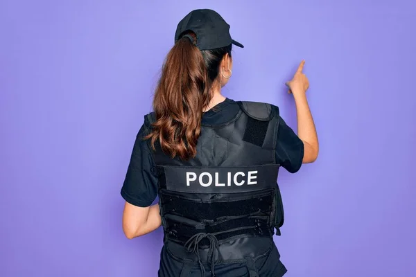 Young police woman wearing security bulletproof vest uniform over purple background Posing backwards pointing ahead with finger hand