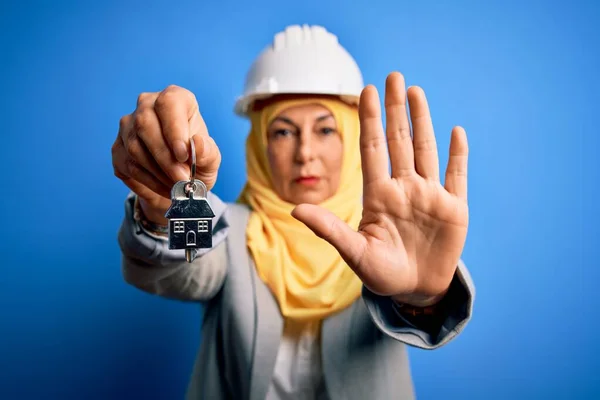 Middle age architect woman wearing muslim hijab and security helmet holding home keys with open hand doing stop sign with serious and confident expression, defense gesture