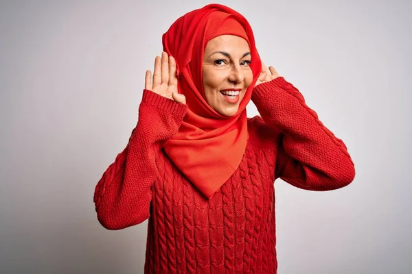 Middle age brunette woman wearing muslim traditional hijab over isolated white background Trying to hear both hands on ear gesture, curious for gossip. Hearing problem, deaf