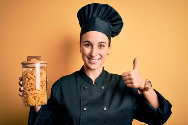 年轻漂亮的厨师女士身穿厨师制服 手里拿着盛有意大利面通心粉的罐子 笑容满面 做手势 用手指竖起大拇指 这是极好的标志 — 图库照片