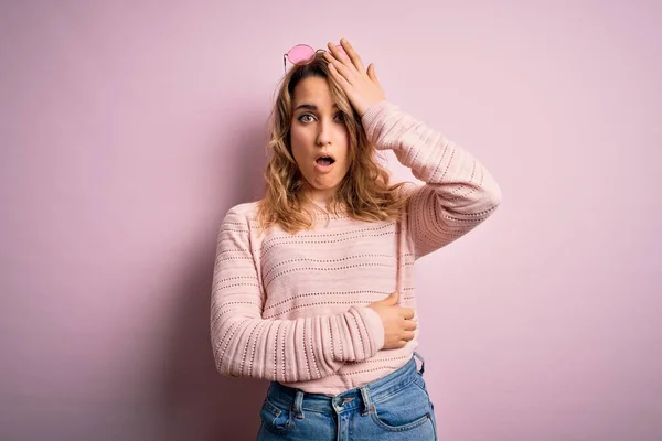 若い美しいブロンドの女性が誤って頭の上に手で驚いてピンクの背景にカジュアルなセーターとサングラスを着て エラーを覚えています 忘れられた悪い記憶の概念 — ストック写真