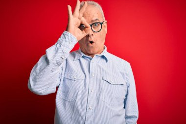 Orta yaşlı yakışıklı beyaz bir adam kırmızı arka planda günlük çizgili gömlek ve gözlük takıyor. Şaşırmış yüz ifadesiyle şaşırmış, parmaklarından bakan gözler. İnançsız ifade.