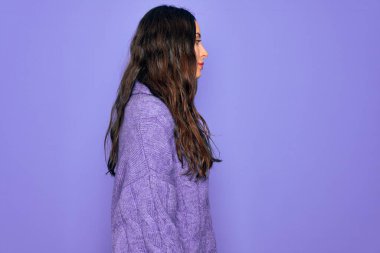 Balıkçı yaka kazak giyen genç ve güzel bir kadın mor arka planda duruyor. Yan yana bakıyor, rahat profil pozu veriyor, doğal bir yüzü ve kendine güvenen bir gülümsemesi var..