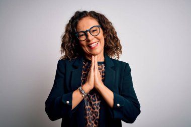 Orta yaşlı esmer iş kadını gözlüklü, izole edilmiş beyaz arka planda durup ellerinin birarada dua ederek af diliyor kendinden emin bir şekilde gülümsüyor..