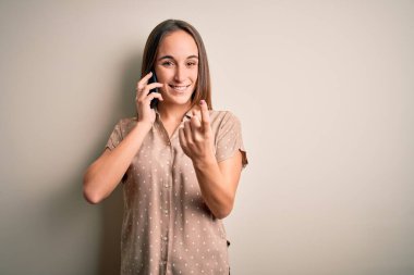 Genç ve güzel bir kadın, beyaz arka planda akıllı telefondan konuşuyor. Buraya gel, el kol hareketi yap, mutlu karşıla ve gülümse.
