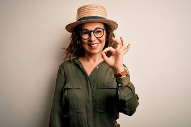 Orta yaşlı esmer kadın, gözlüklü ve şapkalı, izole edilmiş beyaz arka planda duruyor, gülümsüyor ve el ve parmaklarıyla tamam işareti yapıyor. Başarılı ifade.